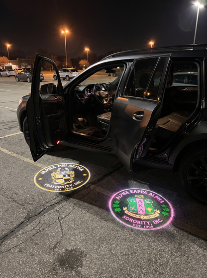 Divine Glow LED car door projectors in use — SUV parked at night with both driver-side doors open, Alpha Phi Alpha and AKA crests projected on the parking lot asphalt