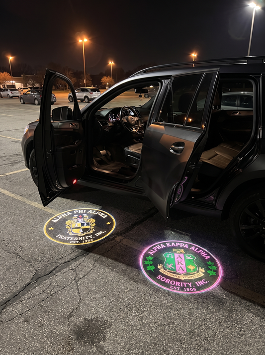 Divine Glow LED car door projectors in use — SUV parked at night with both driver-side doors open, Alpha Phi Alpha and AKA crests projected on the parking lot asphalt